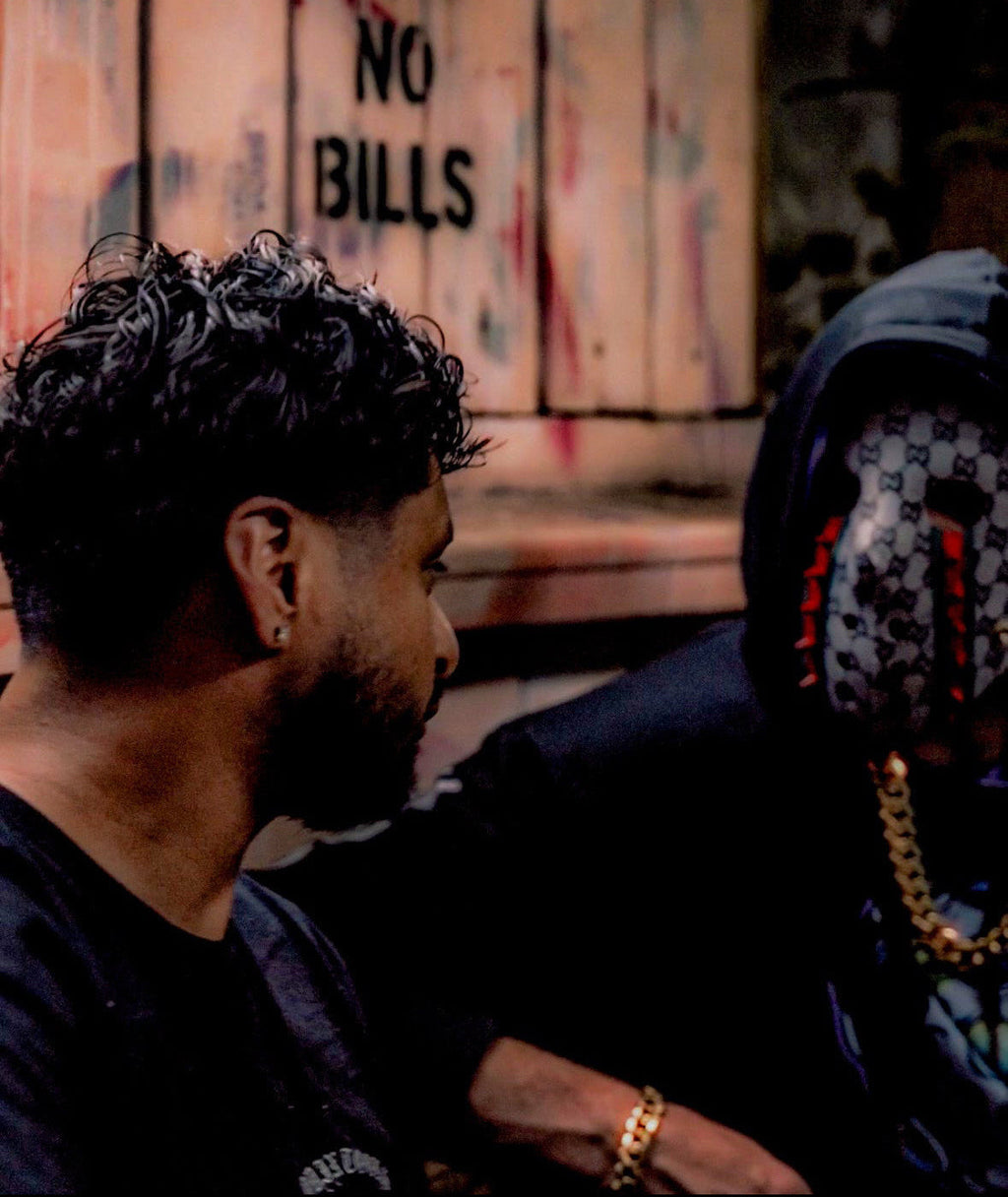 Two people sitting together with a laptop, one wearing a mask, in front of a wall with 'POST NO BILLS' text.